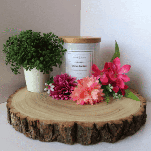 Citrus Garden Candle in white container with wooden lid on, surrounded by flowers and a plant, all sitting on top of a wooden board.