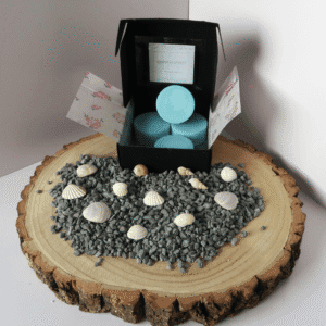 Coastal Walks Wax Melt inside an open black box, surrounded by grey pebbles and shells, all sitting on top of a wooden board.