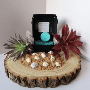 Ho'Omaluhia Wax Melt inside an open black box, surrounded by sand, shells, and plants, all sitting on top of a wooden board.