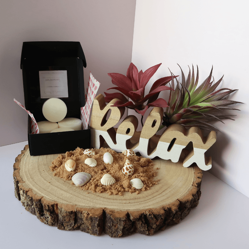Ho'Onanea Wax Melt inside an open black box, surrounded by sand, shells, plants, and a 'relax' sign, all sitting on top of a wooden board.