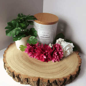 Lavish Embrace Candle in white container with wooden lid on, surrounded by flowers and a plant, all sitting on top of a wooden board.