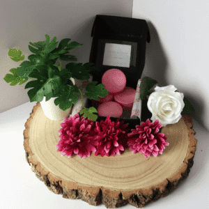 Lavish Embrace Wax Melt inside an open black box, surrounded by flowers and a plant, all sitting on top of a wooden board.