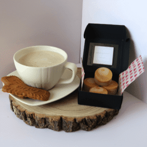 Mocha Madness Wax Melt inside an open black box, beside a cup containing a latte and 2 biscuits on the saucer, all sitting on top of a wooden board.