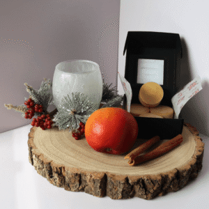 Spiced Orchard Wax Melt inside an open black box, surrounded by a red apple, cinnamon sticks, a red berry and pine needle plants and frosted tealight holer, all sitting on top of a wooden board.