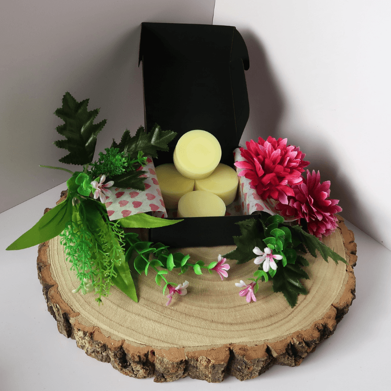 Spring Meadow Wax Melt inside an open black box, surrounded by flowers and plants, all sitting on top of a wooden board.