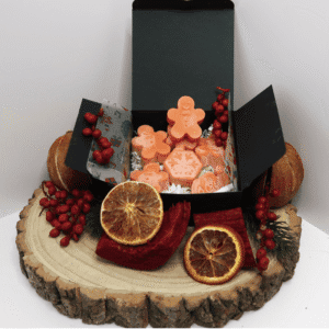 Citrus Spice Wax Melt inside an open black box, surrounded by 2 dried oranges, orange slices, red bark, and red berries, all sitting on top of a wooden board.