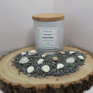 Coastal Walks Candle in white container with wooden lid on, surrounded by grey pebbles and shells, all sitting on top of a wooden board.