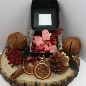 Festive Spice Wax Melt inside an open black box, surrounded by 2 dried oranges, orange slices, gold coloured pinecone, and cinnamon sticks, all sitting on top of a wooden board.