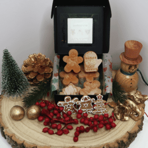 Gingerbread Wax Melt inside an open black box, surrounded by a fir tree, 2 gold balls, red berries, a wooden snowman and gingerbread shapes, all sitting on top of a wooden board.