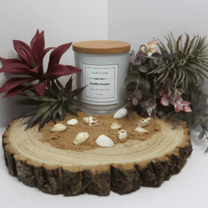 Pacific Fusion Candle in white container with wooden lid on, surrounded by sand, shells, and plants, all sitting on top of a wooden board.