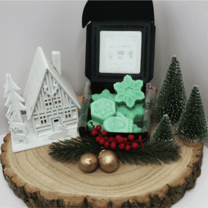 Tis The Season Wax Melt inside an open black box, surrounded by a white wooden house, fir trees, 2 gold balls, and red berries on top of fir branches, all sitting on top of a wooden board.